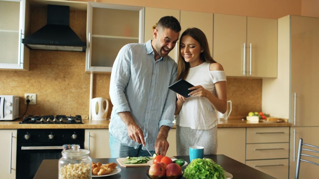 Smiling couple preparing a meal, chopping vegetables and using a tablet in a cozy kitchen setting.