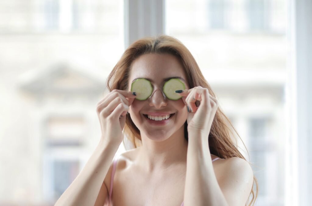 Smiling woman with cucumber slices on eyes, enjoying skincare routine indoors.