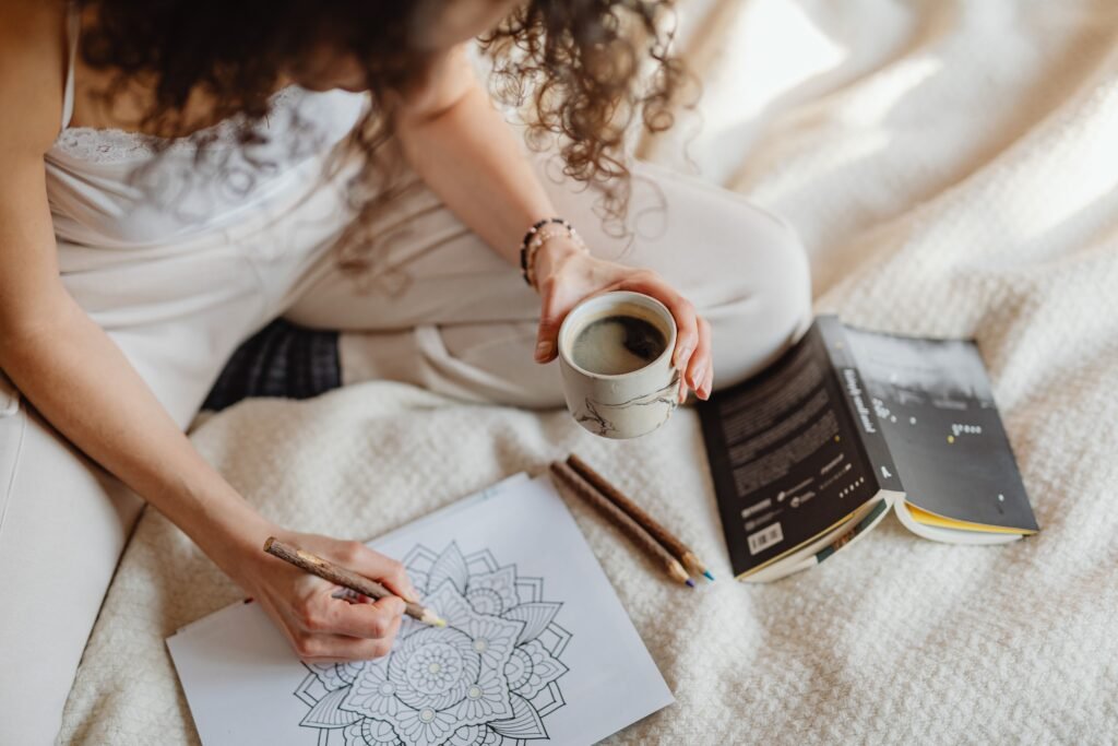 Woman coloring mandala while holding coffee on cozy blanket, enjoying a relaxing moment.