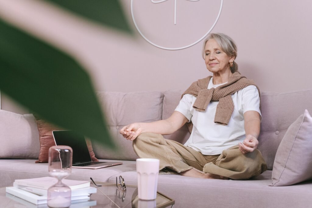 Elderly woman sitting on a sofa meditating with eyes closed, in a peaceful home setting.