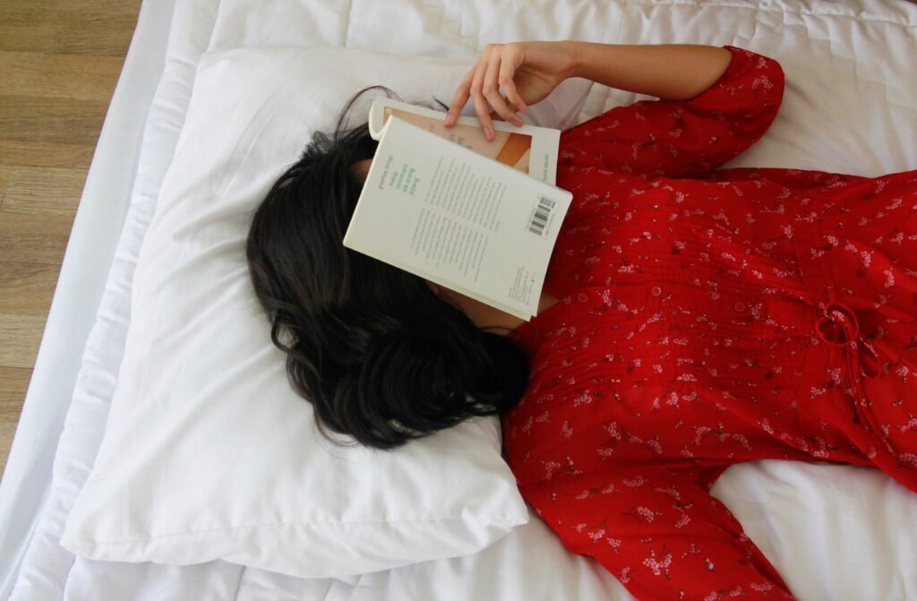 A woman in a red dress lies on a bed with a book on her face, symbolizing relaxation.