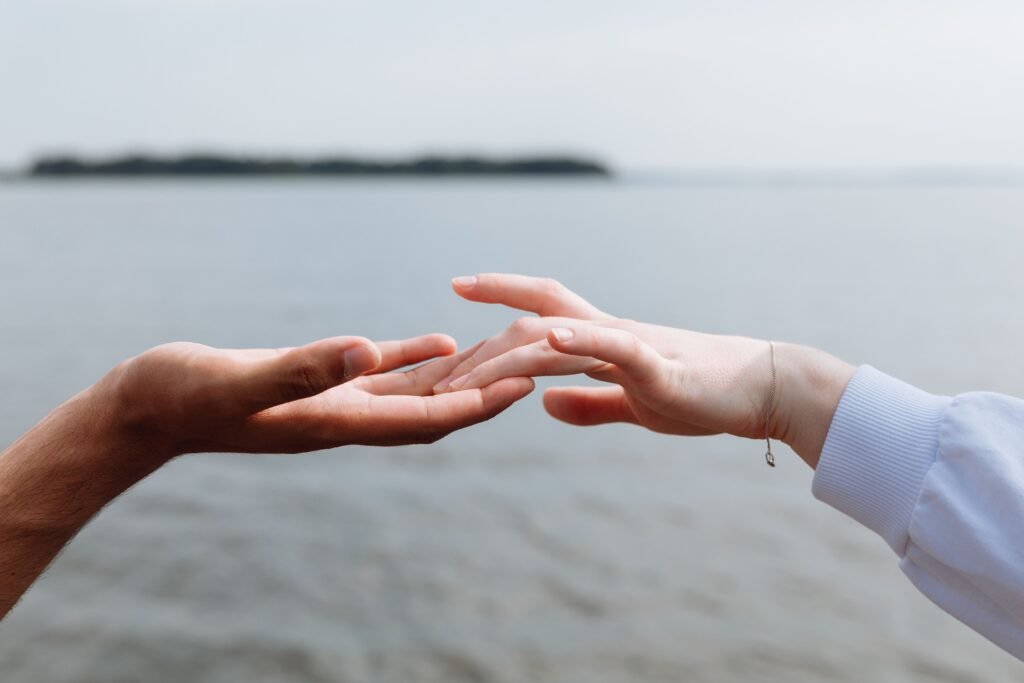 A tender and romantic close-up of two hands gently reaching towards each other against a serene sea backdrop.
