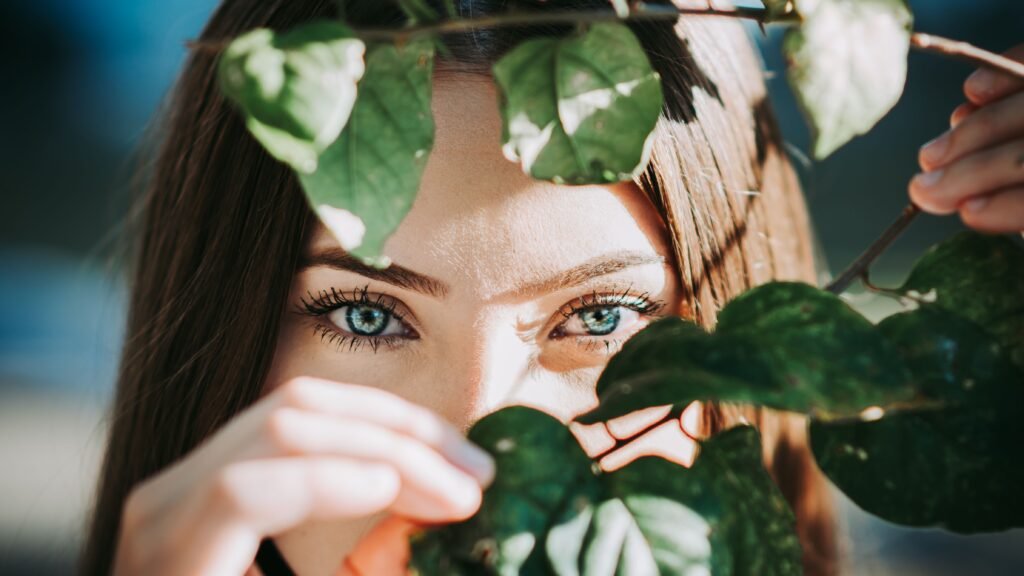 Captivating close-up of a woman with stunning blue eyes peering through vibrant green leaves.