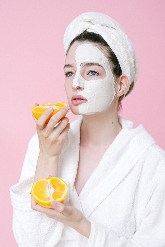 Calm female with towel on head and facial mask applied on half face looking away while standing on pink background with halves of oranges in hands