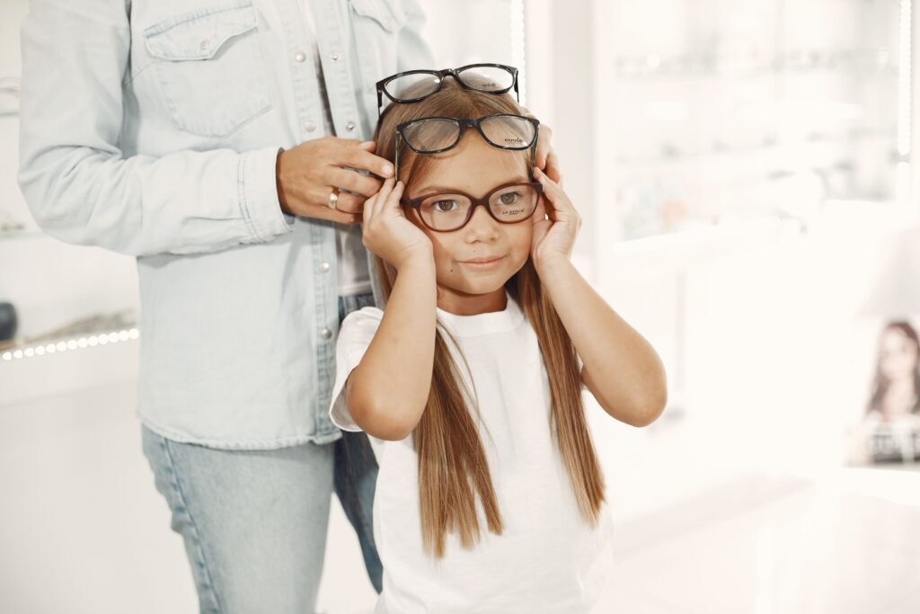 Young girl in a white shirt trying on glasses indoors, assisted by an adult.