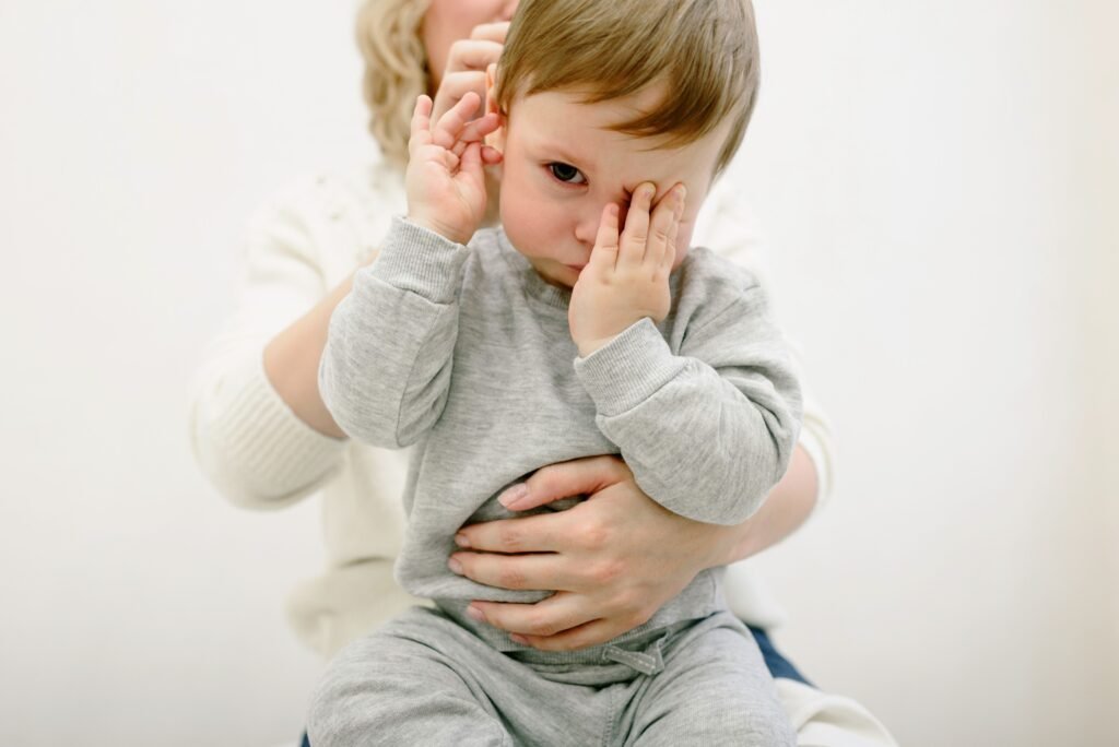 Cute baby in grey, held by adult, covering eyes playfully. Warm and cozy indoor setting.